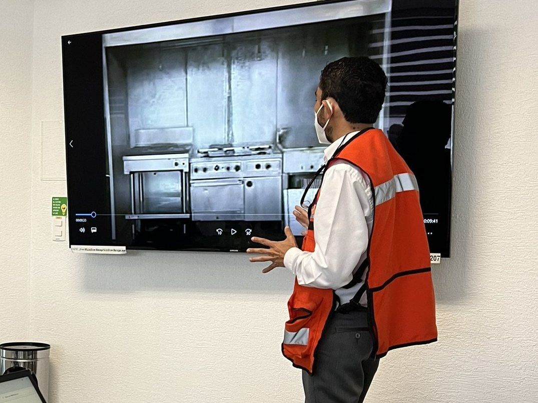 Especialista con chaleco de alta visibilidad presenta en una pantalla de pared el video de una cocina industrial.