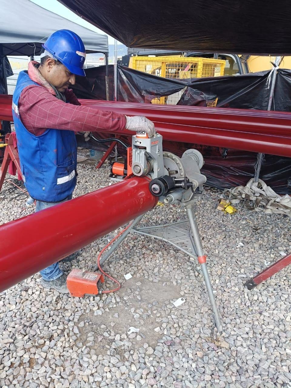 Técnico ranura un tramo de tubería roja de acero con una ranuradora montada en su tripié, en el patio de una obra.