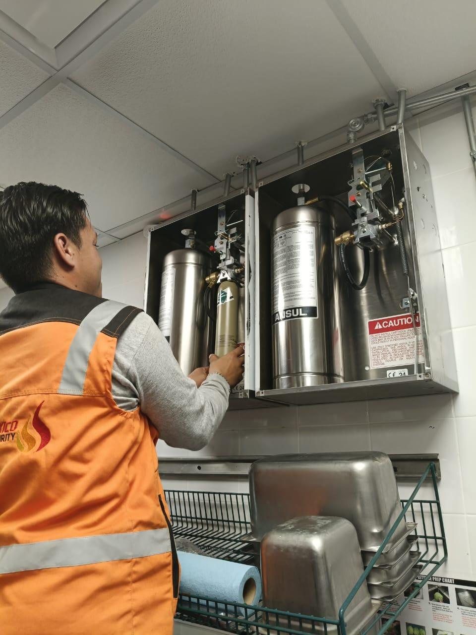 Técnico con chaleco revisa el gabinete abierto de un sistema Ansul con dos tanques de agente, en una cocina industrial.
